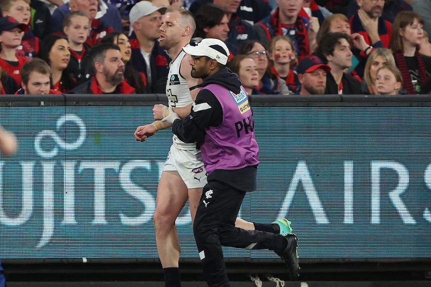 A Carlton AFL player grimaces as he is helped off the ground with a dislocated shoulder.