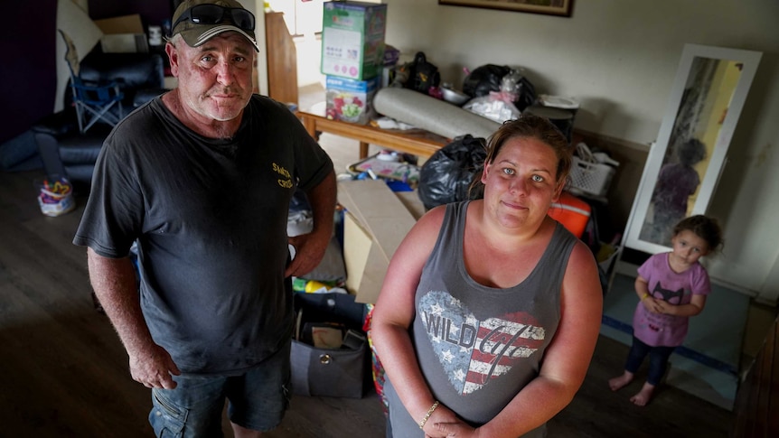 Man with cap, t-shirt and shorts and woman in singlet stand with small girl behind them in cluttered room