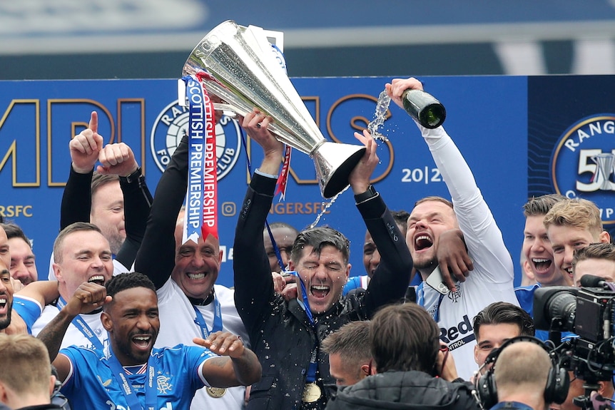 Rangers players celebrates with manager Steven Gerrard, who's lifting a trophy and having champagne poured on his head.