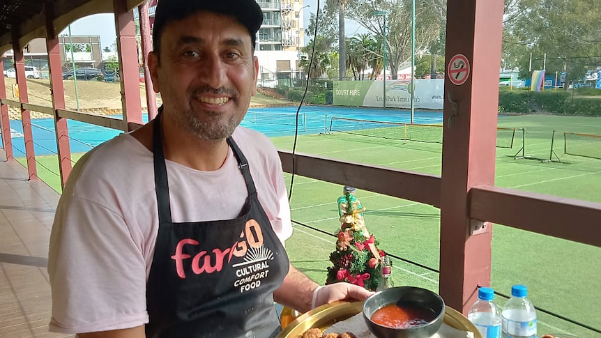 A smiling man wearing an apron holds a plate of falafel balls on the veranda of a tennis club.