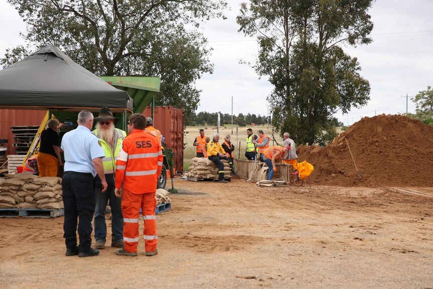 Three people, including a man in a high-visibility SES jumpsuit, stand near a sandbagging facility