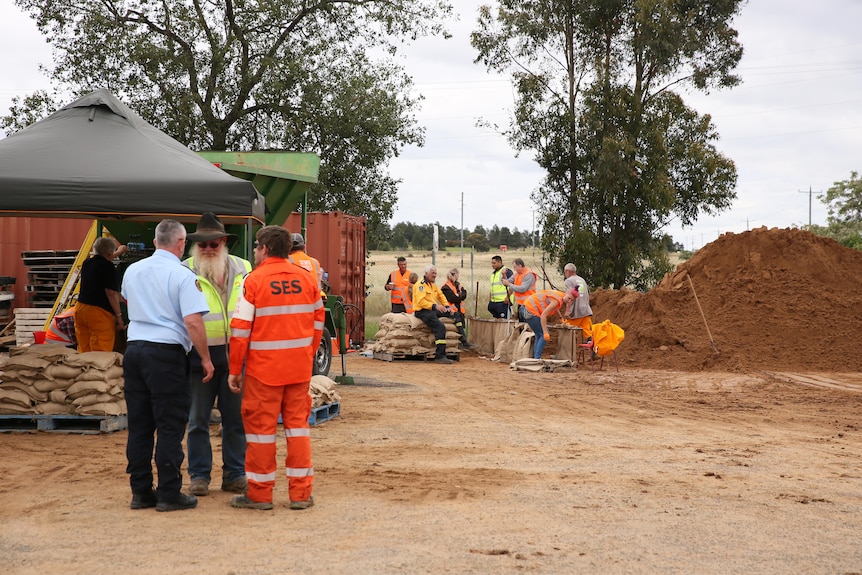 Three people, including a man in a high-visibility SES jumpsuit, stand near a sandbagging facility