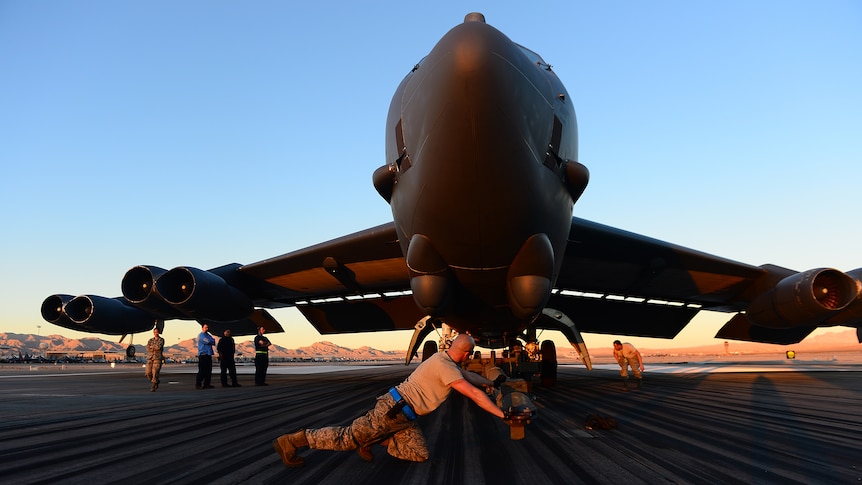 A photo taken looking up at a massive military aircraft. A soldier is pushing a tow bar towards its front wheel.
