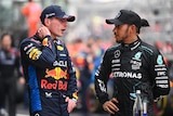 F1 drivers Max Vertsappen and Lewis Hamilton, in racing suits with no helmet, speak to each other in the pit lane