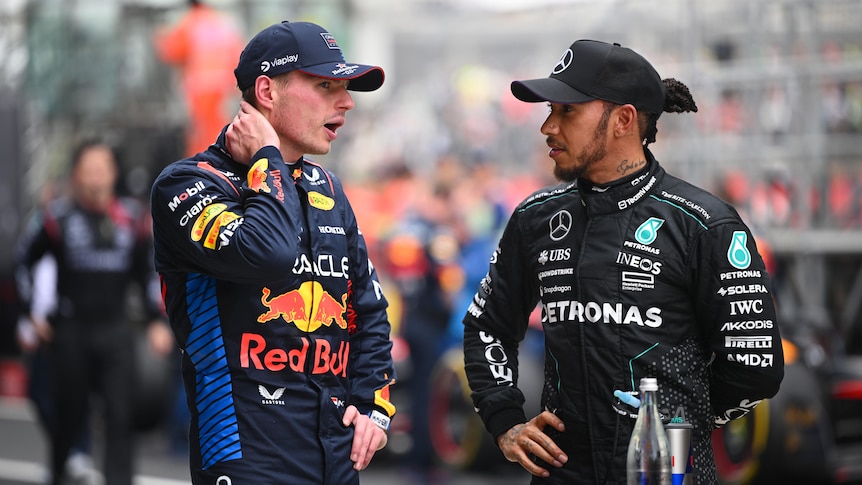 F1 drivers Max Vertsappen and Lewis Hamilton, in racing suits with no helmet, speak to each other in the pit lane