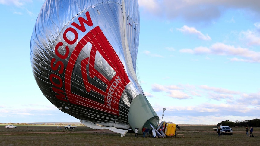 Large silver hot air balloon lists to one side after touching down in a grassy paddock.