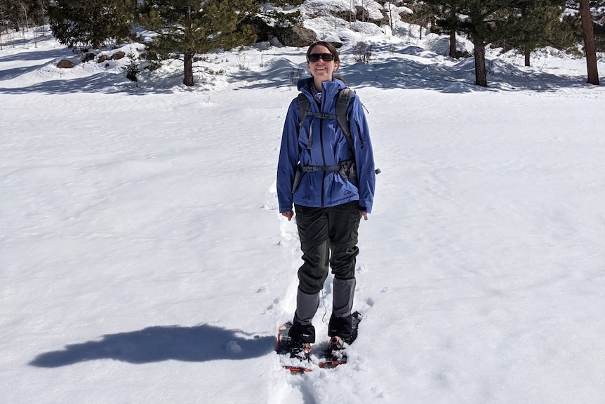 A woman in snow attire standing in fresh snow with a forest behind.