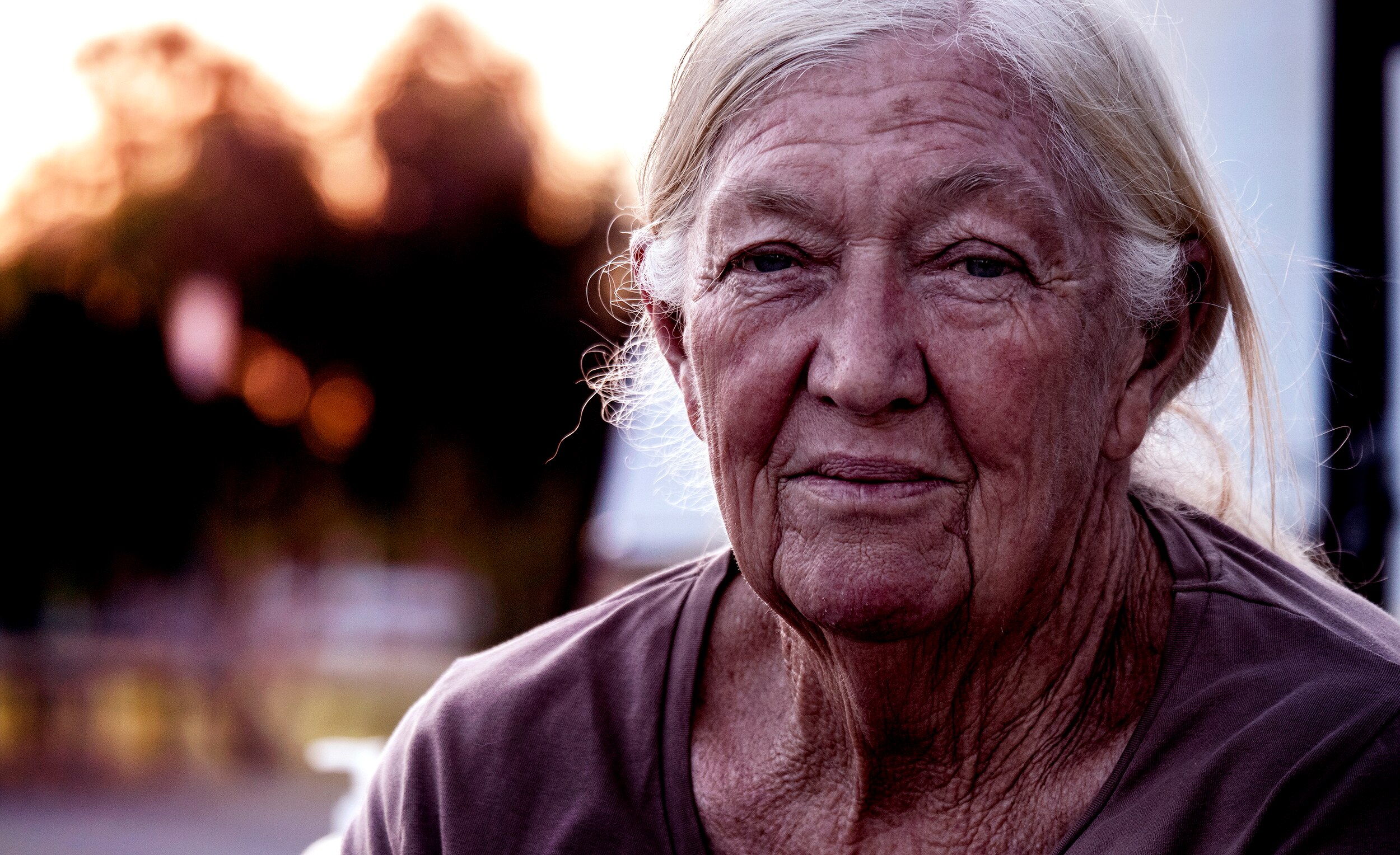 A close up photo of an elderly women with a weathered face, grey hair, blurred trees in background.