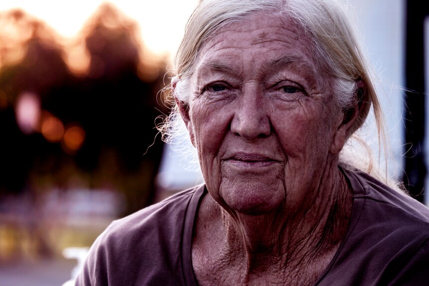 A close up photo of an elderly women with a weathered face, grey hair, blurred trees in background.