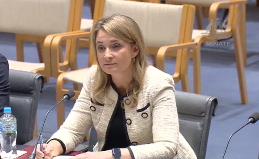 A blonde middle-aged woman in a suit sits in a grey chair in front of a microphone.