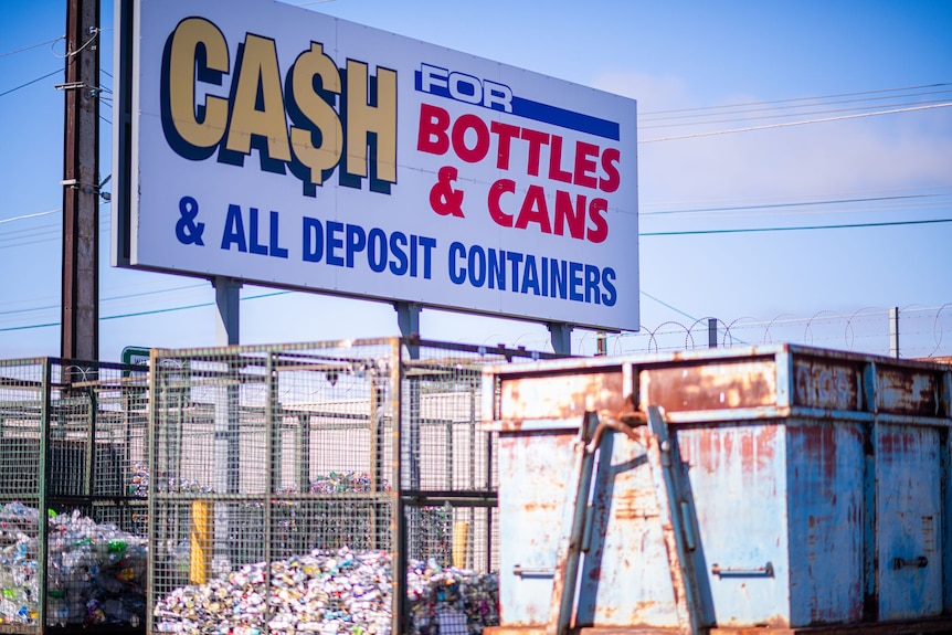 A large sign that says 'cash for bottles and cans' with recycled cans and bottles in crates in the background. 