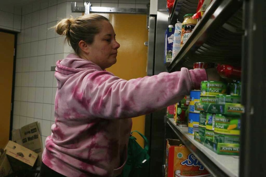 A woman in a pink jumper reaches for a can of food that's sitting on a shelf