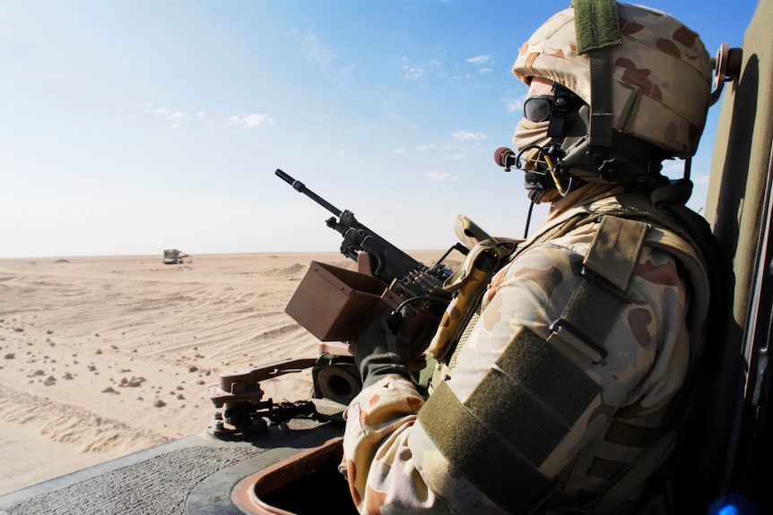 A soldier in camouflage with a helmet and face covering sits in the back of a bushmaster with a gun looking over a desert