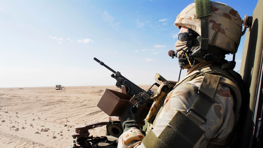 A soldier in camouflage with a helmet and face covering sits in the back of a bushmaster with a gun looking over a desert