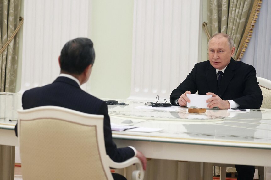 Putin sits at an ornate white table, a small earpeice in his ear. Opposite him is Wang Yi, who we see from the back