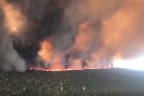 An aerial photo of a long fire front burning in bushland sending large dark clouds of smoke into the air.