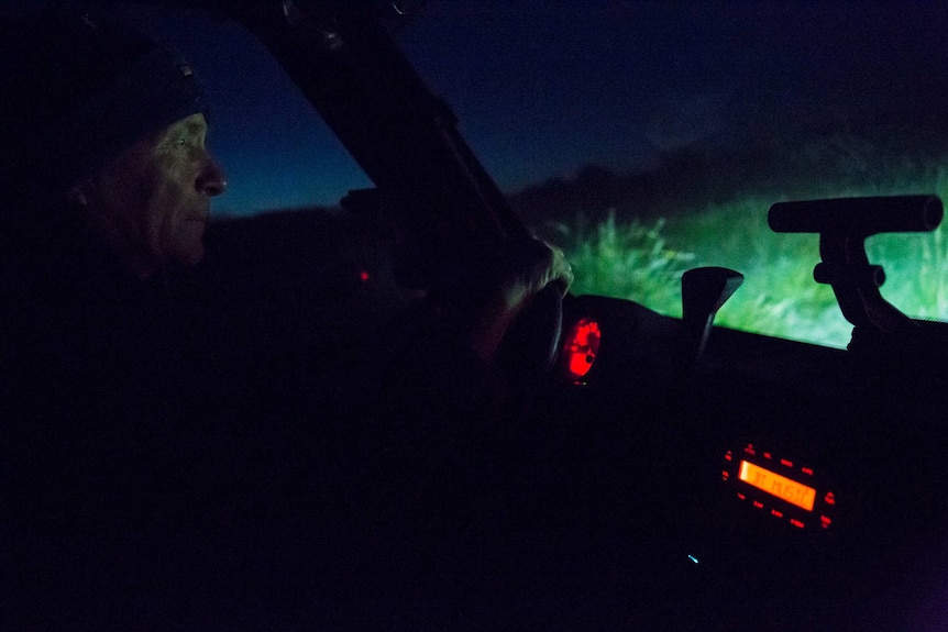 Tony Symes' face is lit by the red lights of the dash in his buggy, the sky and grass glowing blue and green outside.