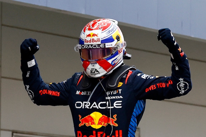 Max Verstappen, in his racing suit and helmet, raises his fists in the air in celebration after winning the race.