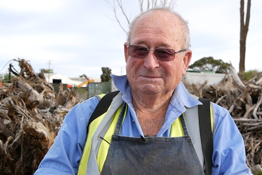 He wears an apron and stands near a pile of logs