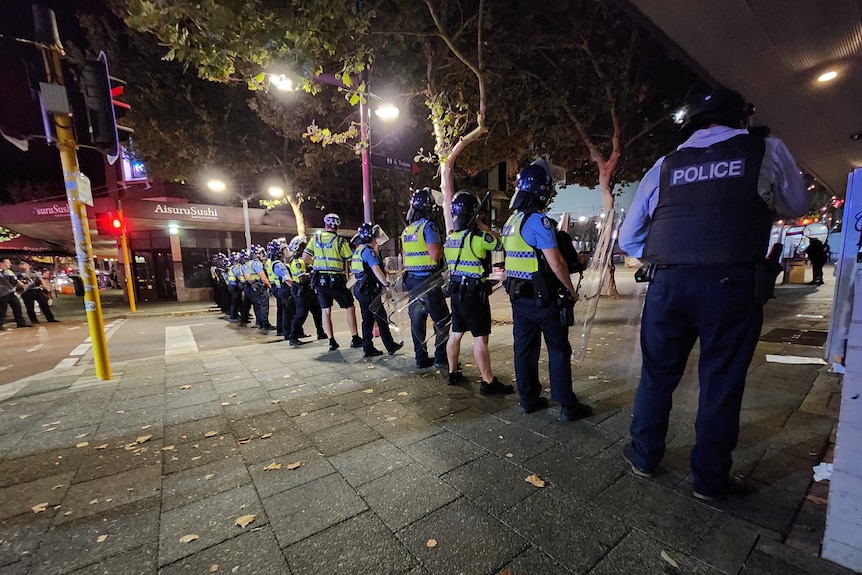 A line of police in riot gear stands across a road in Perth's CBD at night.