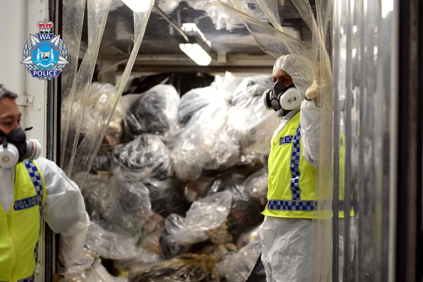 police in forensic suits and masks in front of a pile of plastic sealed rubbish bags.