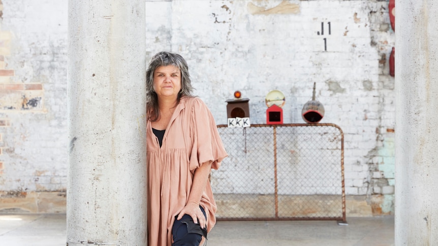 A 50-something Aboriginal woman with a mullet crouches beside a white pole; behind her are letterboxes on a gate