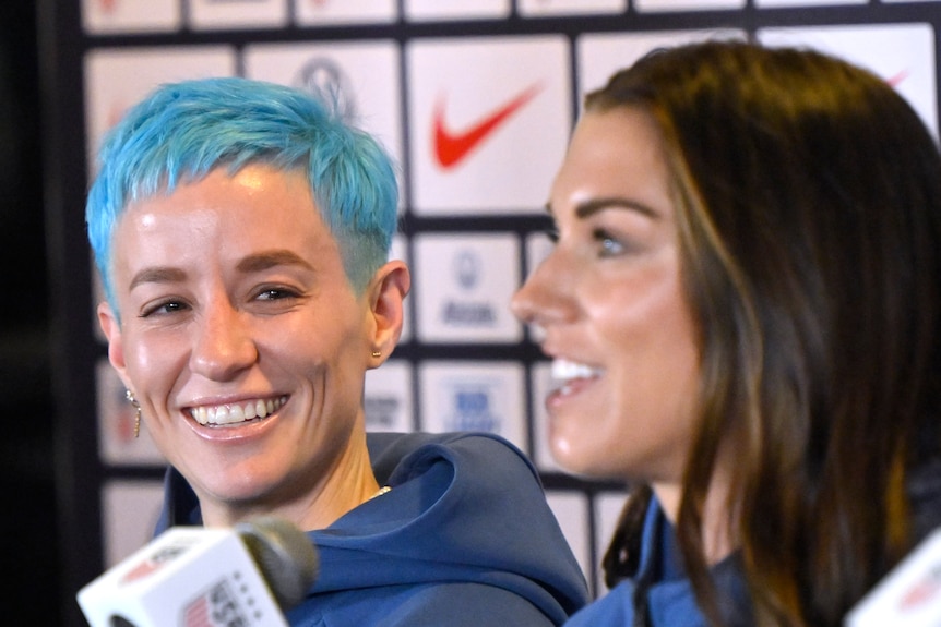 A football player with blue hair smiles at another player as the take questions at a press conference.