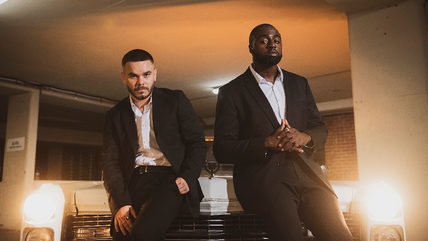 Two men, both dressed in black suits with white shirts lean against car with lights on