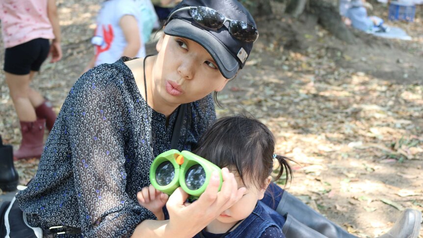 A mum wearing a hat holds up binoculars for her toddler sitting on a mat in the forest