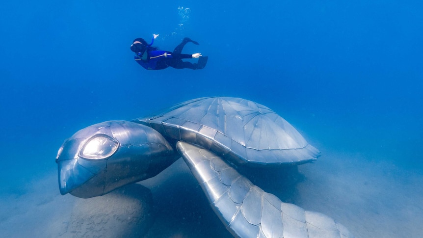 A large metal sculpture of a turtle lays on the sand underwater, as a diver swims above.