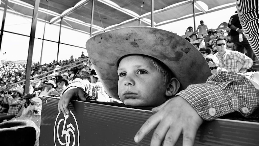 A black and white photo of a young boy leaning on the railing of a rodeo, with a crowd of people seated behind him.