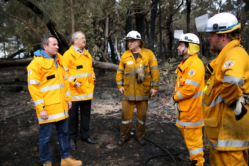 Firefighters and the premier stand in full protective gear in burnt out bushland.