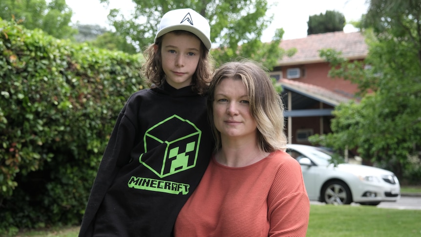 A young boy and a woman standing close together, with a house in the background