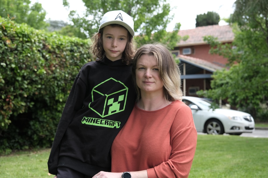 A young boy and a woman standing close together, with a house in the background