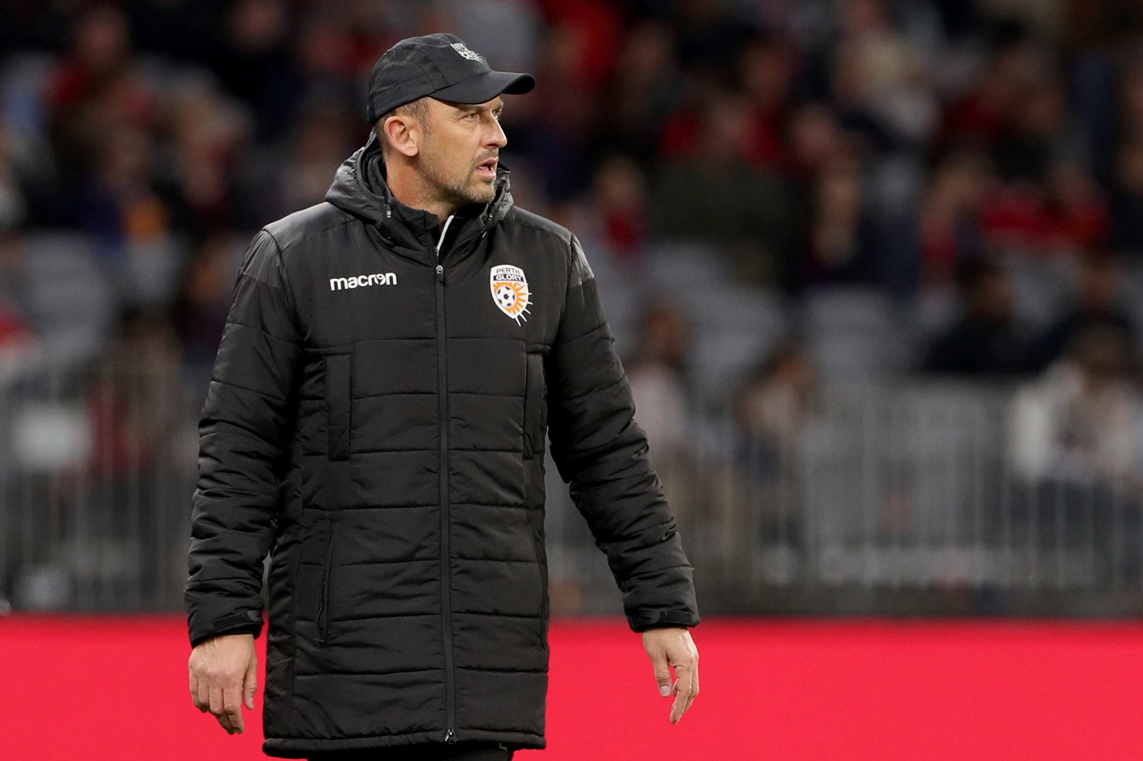 A mid shot of Perth Glory coach Tony Popovic on the sidelines wearing a black winter jacket and baseball cap at night.
