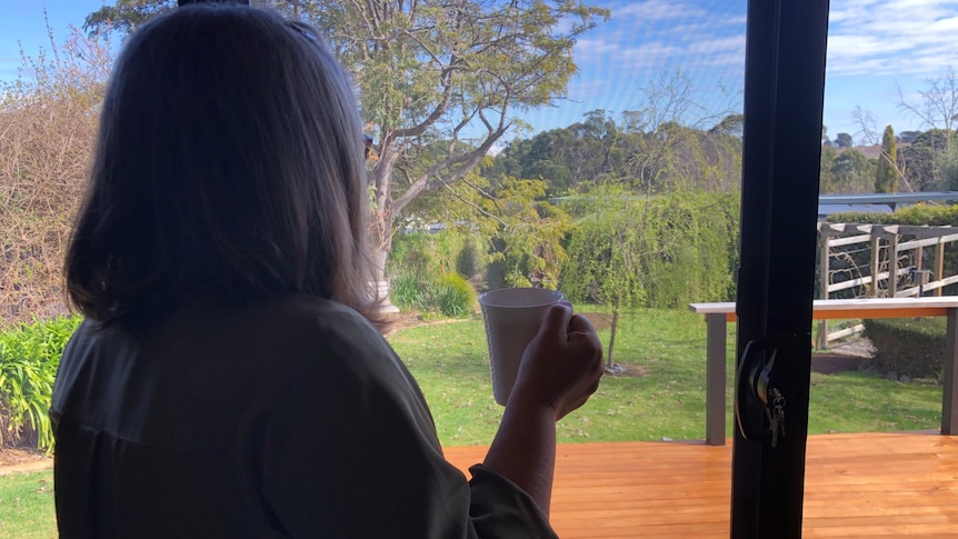 A woman stands with her back to the camera, holding a cup of tea, looking out the window