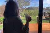 A woman stands with her back to the camera, holding a cup of tea, looking out the window