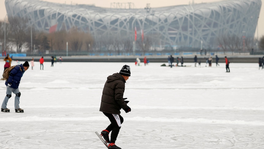 A man dressed in black skates on ice with a big stadium in the distance.