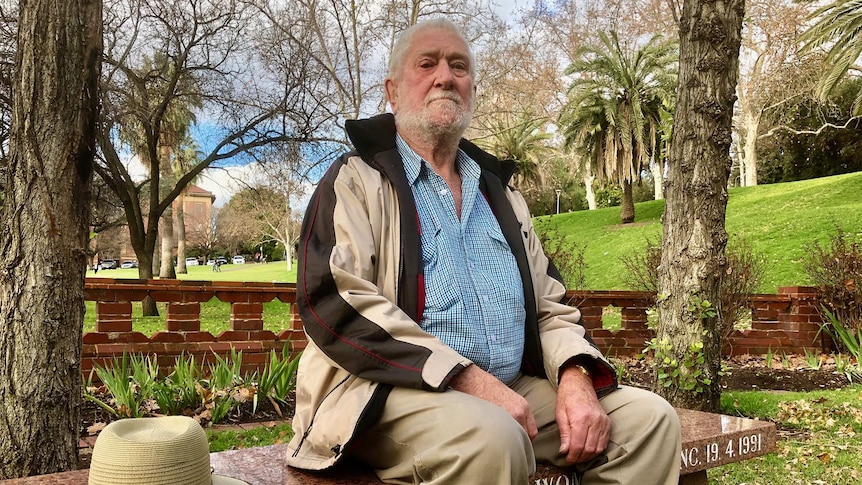 A wide shot of Ian Stewart posing for a photo sitting on a park bench outdoors with his hat alongside him.