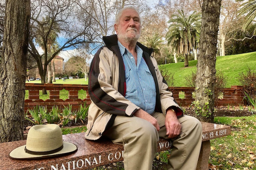 A wide shot of Ian Stewart posing for a photo sitting on a park bench outdoors with his hat alongside him.