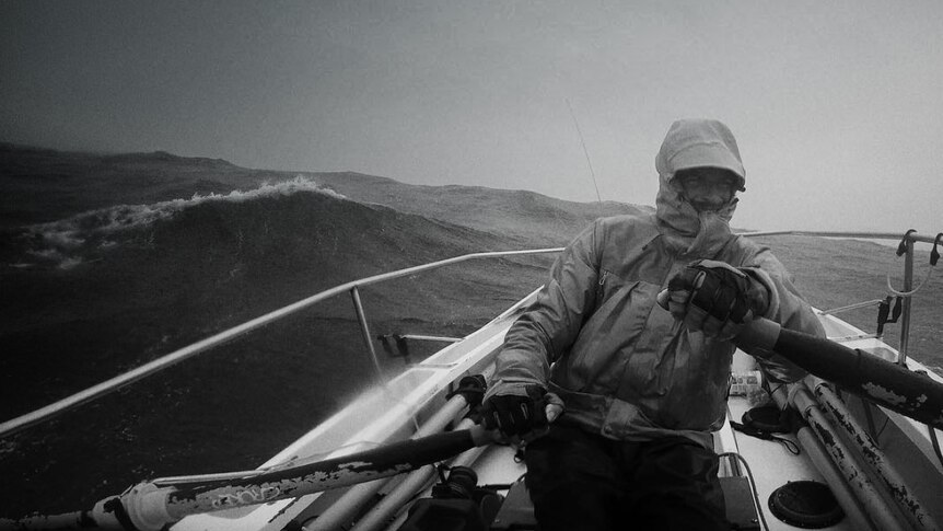 A black and white photo of a man rugged up in wet weather clothes rowing across the ocean.