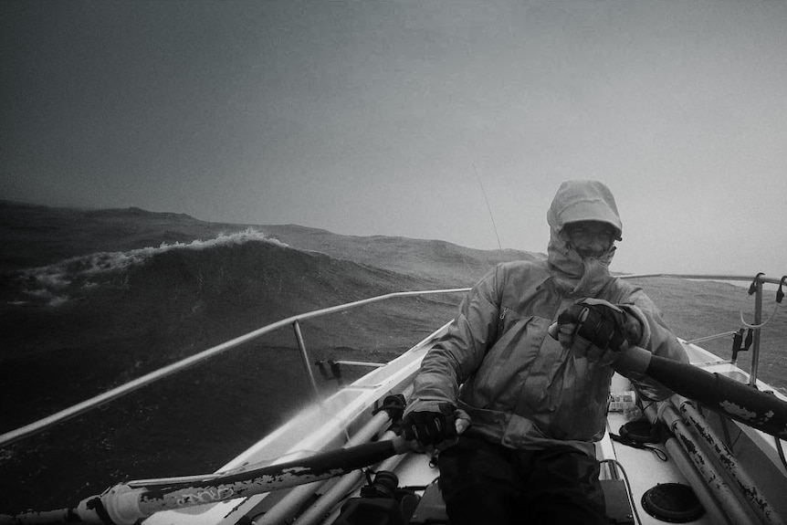 A black and white photo of a man rugged up in wet weather clothes rowing across the ocean.