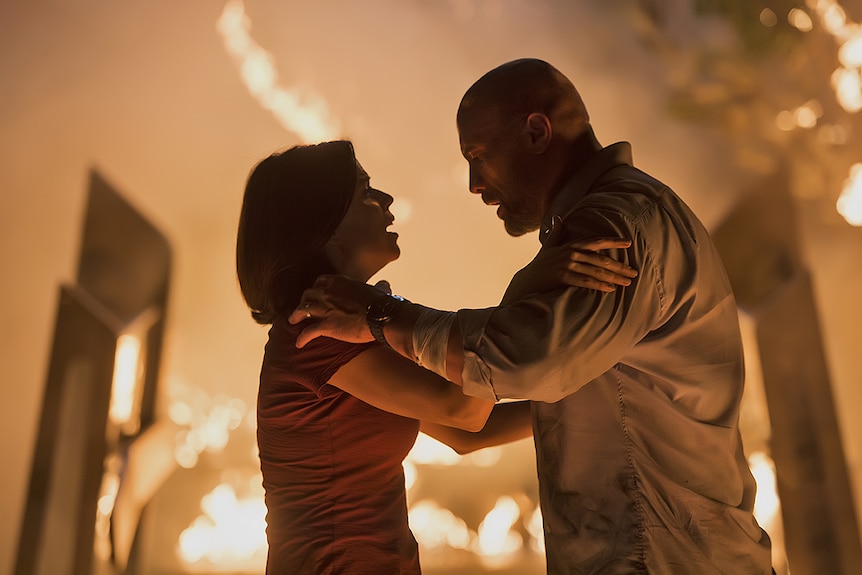 Colour still image of Dwayne Johnson and Neve Campbell holding each other in front of a fiery backdrop.