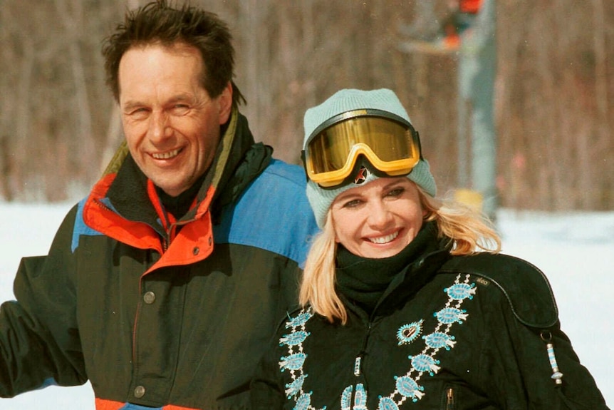 A man in ski gear stands just behind a blonde woman who has ski goggles perched on her beanie. They are in a snowfield