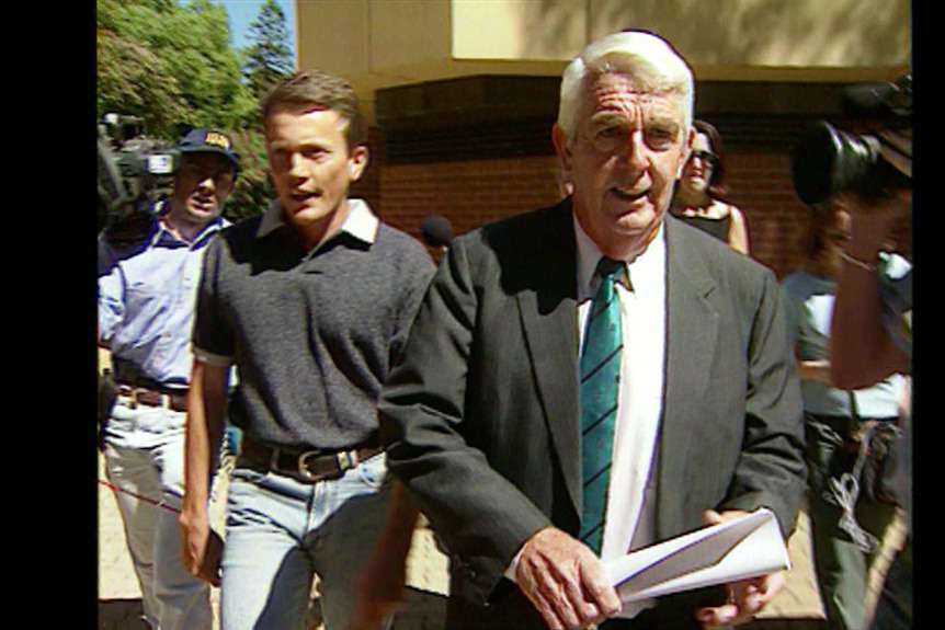 Two men walking surrounded by TV cameras and journalists