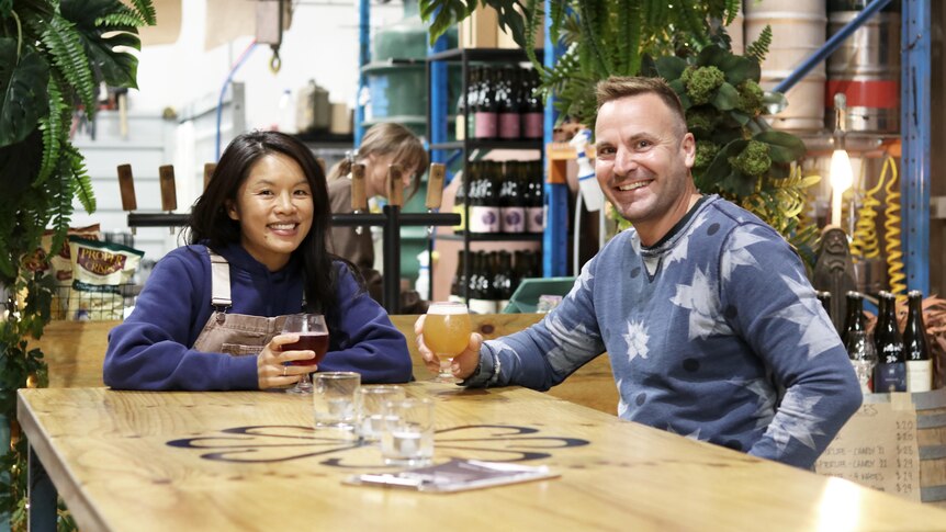 Chen and Georg sit at a wooden table and smile while holding glasses of beer with the bar behind them.