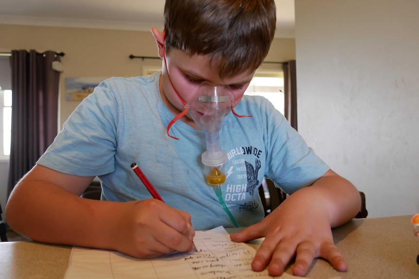 Lincon sits at kitchen counter with breathing mask over his face, leaning over doing homework.