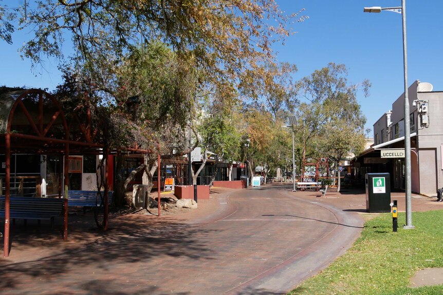 The glare of the sun hits the bitumen of an Alice Springs street lined with shops and trees.