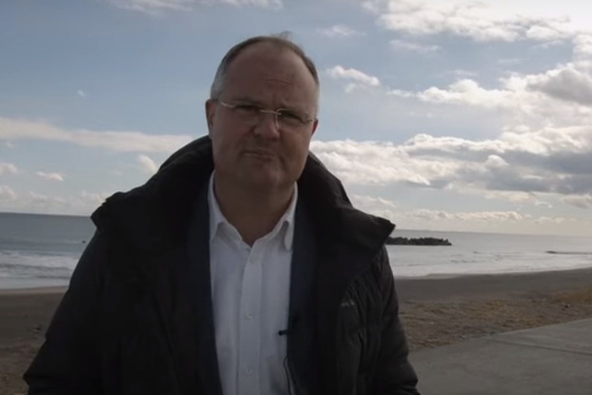 A bald man in a puffer jacket and business shirt stands on a beach looking into a phone camera