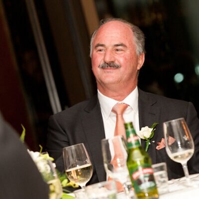 A man in suit and tie sits at a table with glasses of wine and bottles of beer on it.